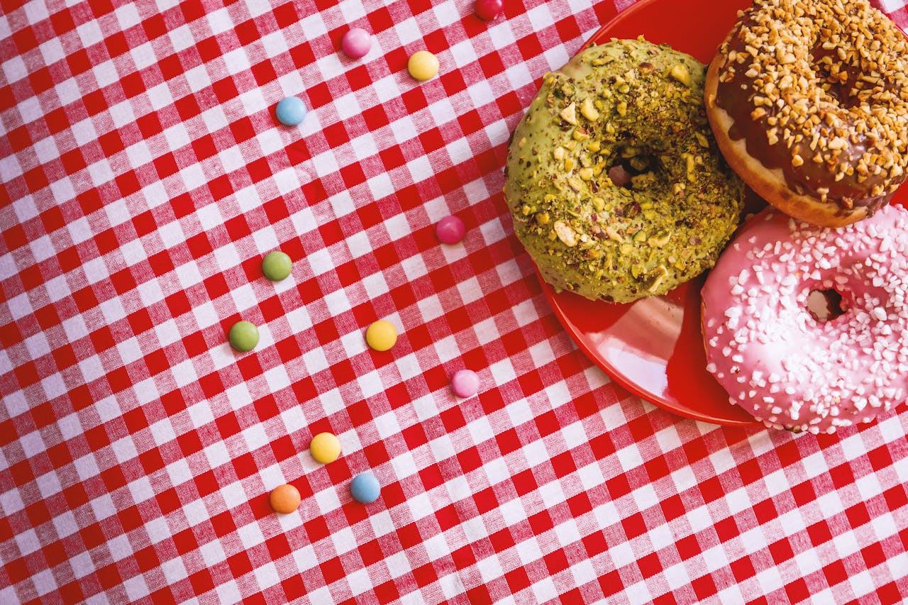 Three decorated donuts on a red checkered tablecloth with colorful candies scattered.
